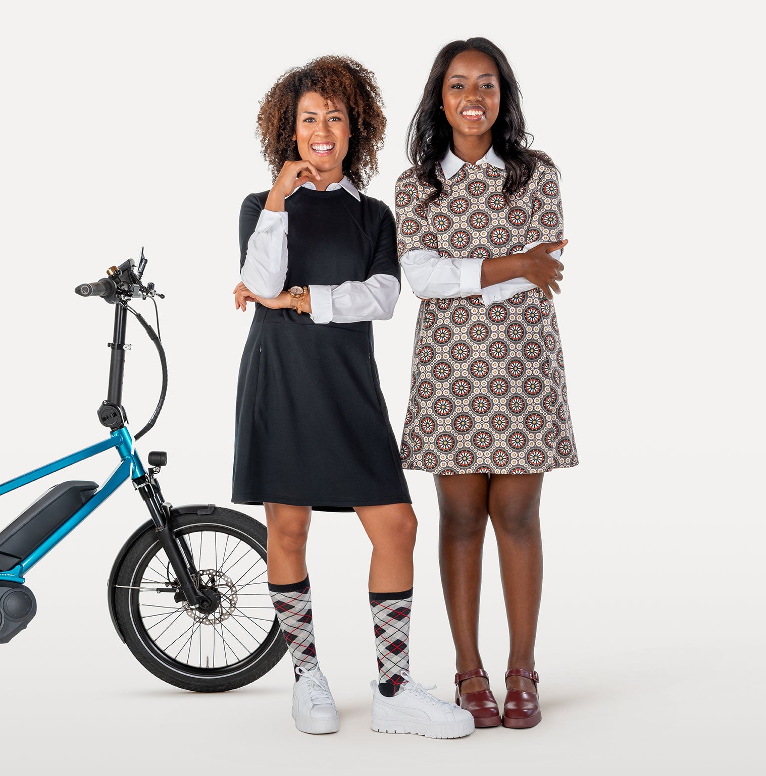 Two women in biking dresses next to a blue electric bike on a white background