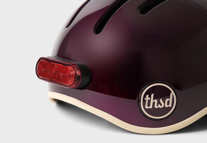 Close-up of a dark purple helmet with a red light and 'thsd' logo on a light gray background
