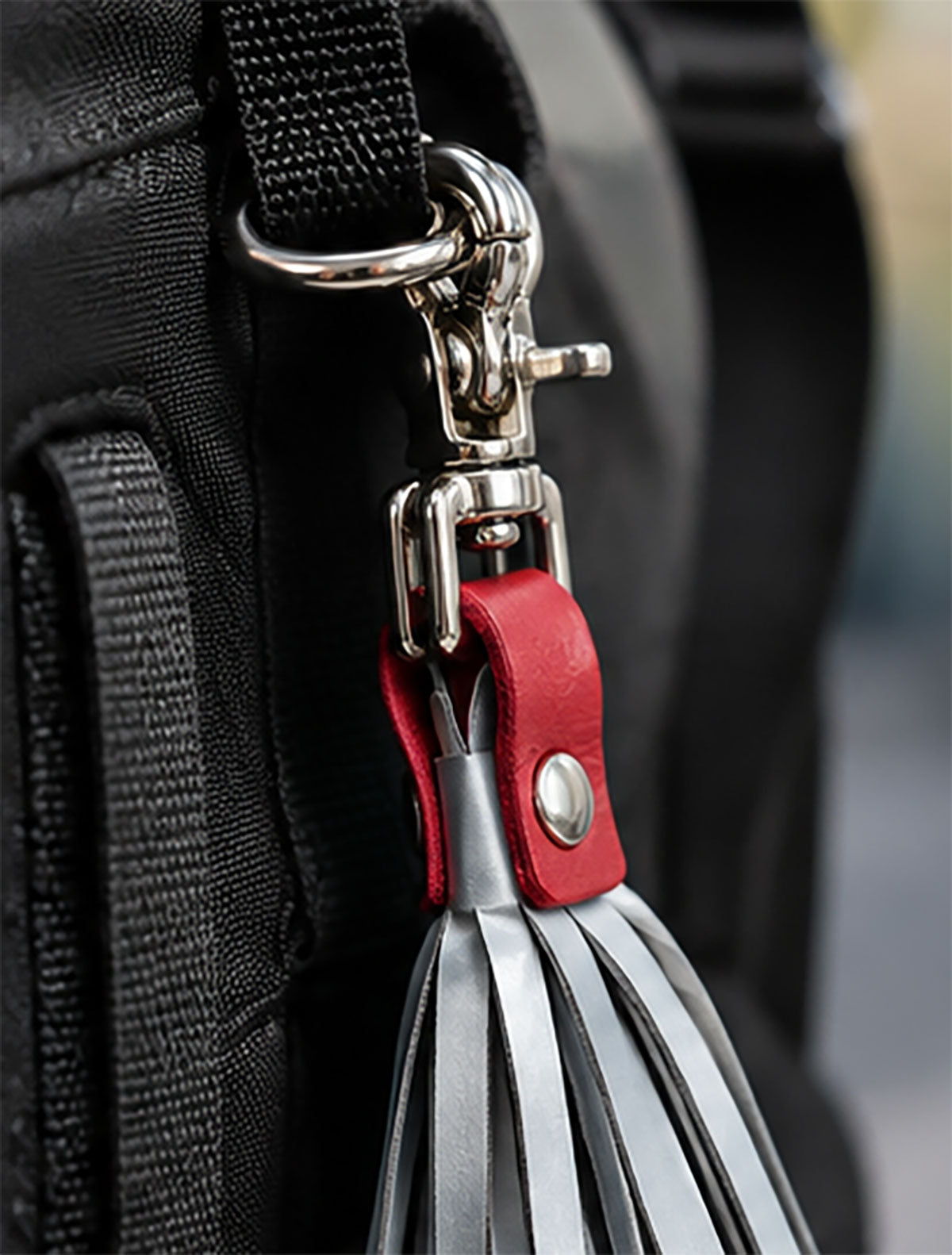 Close-up of a black bag strap attachment with a silver metal clasp and a red leather loop holding grouped silver tassels.