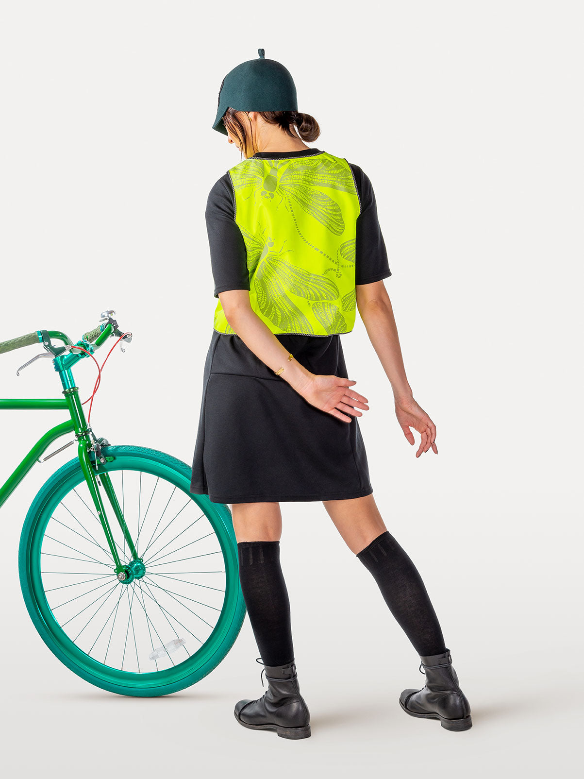 Person wearing a reflective high-vis vest and woolen cap standing next to a green bicycle on a white background